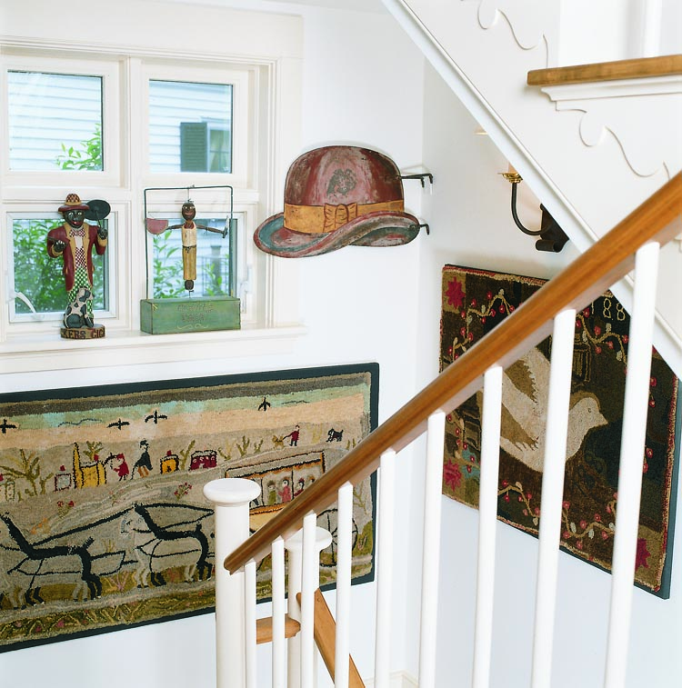 Decorating the lower stair landing is an 1887 Lancaster, Pennsylvania, hooked rug from Olde Hope Antiques, and a 1910 rug with a horse and carriage scene from Jeff Bridgman. The 19th-century hat trade sign from the Dr. and Mrs. Ed Hoffman collection was purchased at Northeast Auctions, Portsmouth, New Hampshire. On the window sill is a 20th-century figure by outsider artist William Duffy (left), and an 1880s figure of a man slicing a watermelon.