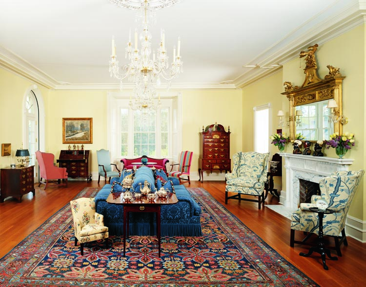 “We lean toward Massachusetts and Rhode Island furniture of the Queen Anne through Federal period,” says the husband. One important piece, rear right, is a carved, walnut-veneered and gilded Boston Queen Anne high chest of drawers. John Kirk cites the casepiece, one of five similar examples known, as an outstanding example in his comparative study, American Furniture. Two Chippendale wing chairs flank the fireplace. One, a Boston example, is signed John Cogswell. The other, from Newport, is attributed to John Goddard. Another, in front of the slant-front desk, was originally owned by John Brown of Newport. In the foreground, an inlaid Charleston Pembroke table supports Chinese export silver. The child’s chair, left, in eighteenth-century embroidered fabric is from Cora Ginsburg. The circa 1890 Bidjar carpet is from Taylor B. Williams.