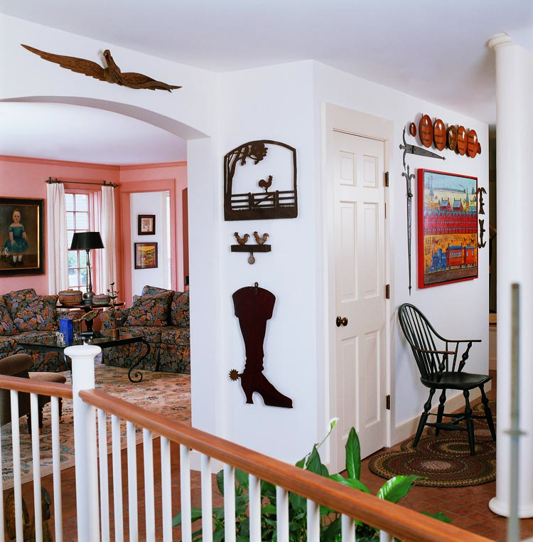 : Hanging in the living room (to the left in the photograph) is a portrait of a young boy in a blue dress attributed by expert Don Walters to William Matthew Prior (1806-1873), circa 1845. Over the doorway is a carved “E. Pluribus Unum” eagle of 1875, attributed to John Haley Bellamy (1836–1914) of Portsmouth, New Hampshire, and Kittery, Maine. At 45 inches long, it has an unusually wide wingspan for a Bellamy. Ironware is mounted on the wall between the living room and hallway: a cast-iron barnyard scene from Pennsylvania hangs above a double rooster shooting target and a shoemaker’s trade sign. A Rhode Island continuous-arm Windsor in the hallway is situated under a monumental Jack Savitsky (1910–1991) painting, Reading Rail Road Miners’ Train, framed by early- 19th-century architectural iron hinges from dealer Rick Jorgensen. Mid- to late-19th-century redware plates from Olde Hope Antiques surmount the painting and hinges.