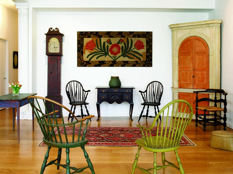The unusual floral border on the boldly patterned New England hooked rug, circa 1840–60, echoes that on a flat-top box in the owners’ collection. The rug came from Elliott and Grace Snyder, who also supplied the circa-1770 North Shore, Massachusetts, dressing table and the 1815 paint-decorated tall-case clock, left, housing works by Lumen Watson of Cincinnati, Ohio. On the dressing table is the circa-1850 New England swing-handle basket that came from Barbara Pollack. The Portsmouth, New Hampshire, architectural corner cupboard of circa 1770–80 and two Rhode Island brace-back Windsor armchairs, flanking the dressing table, are from Nathan Liverant & Son. The Massachusetts Queen Anne corner chair, right, in Spanish brown over original red paint, is ex-collection of Sandy and Julie Palley. In the foreground are two late-18th-century Rhode Island Windsor armchairs.
