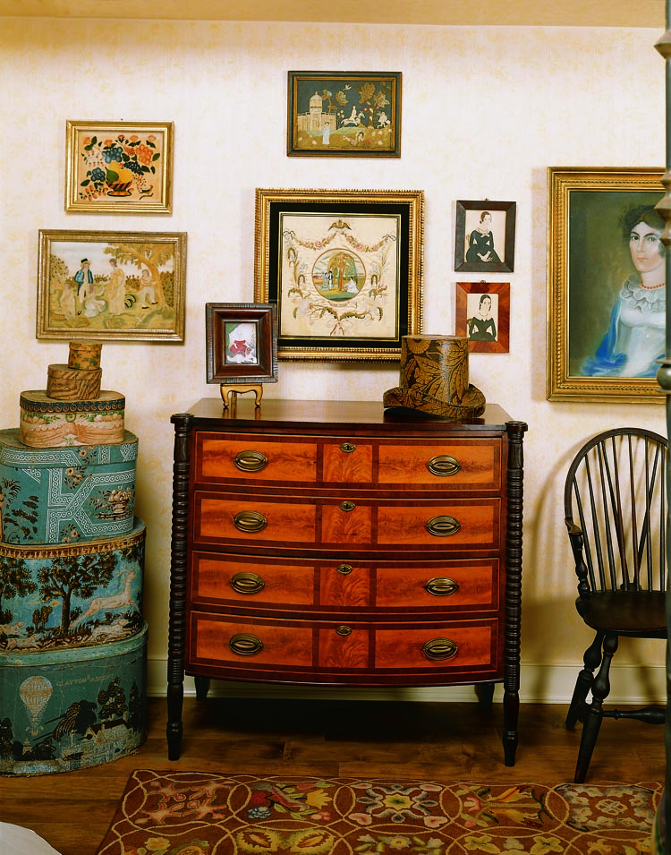 Alternating panels of flame birch outlined by mahogany crossbanding, as present in this chest, are the hallmarks of Federal furniture from New Hampshire. The 19th-century wallpaper-covered bandboxes are just a sampling of what the couple own. The elaborate hat box on top of the chest is from the Miriam and Arthur Spector collection. Also on the chest is a miniature watercolor on ivory of a young child, attributed to Mrs. Moses B. Russell (1809–1854), and purchased from Frank and Barbara Pollack. Needlework, a theorem, watercolors (attributed to J. A. Davis (1821–1855)), and a pastel of Abigail West by William M. S. Doyle (1769–1828) are displayed on the wall.