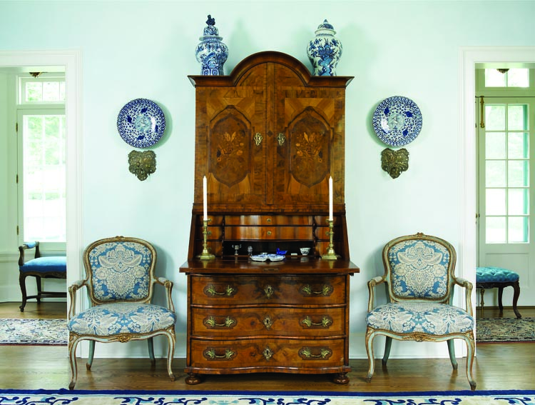 A finely inlaid early eighteenth-century Northern European secretary is flanked by a symmetrical arrangement of eighteenth-century French side chairs and pieces of Dutch Delft ceramics. Beyond, is a hall gallery where south-facing floor-to-ceiling French doors provide views of the garden.
