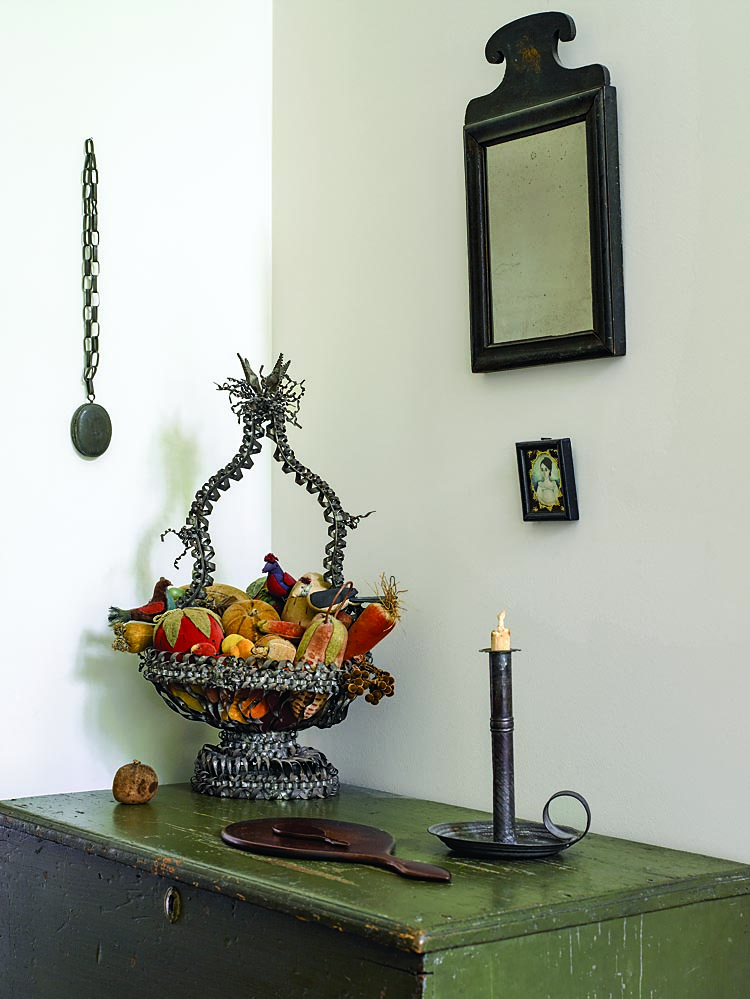 This corner of the bedroom holds a collection of “anniversary” tin. The tradition of making or giving tenth wedding anniversary gifts out of tin was prevalent in the late nineteenth century, particularly in the 1870s–1880s. The elaborate basket filled with velvet fruit (ca. 1840–1870) was in the collection of Barry Cohen; the owner has retained the grouping exactly as Cohen displayed it. “This is one of my most favorite pieces,” says the owner, “I’ve never seen a comparable example.” the other tin items displayed here include a lady’s locket hanging on the wall, a very early wedding-band candlestick, and a hand-held mirror backed with tin. Seen also is a portrait miniature on ivory and an early Queen Ann japanned mirror with gilt trim.