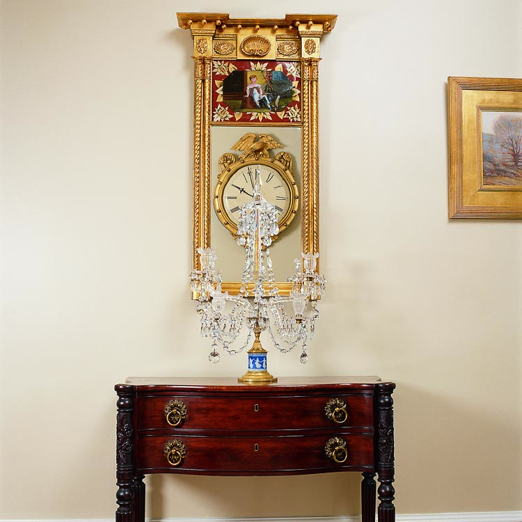 The rose and star-punched caving on this circa 1810–1815 Federal two-drawer, serpentine-front serving table, possibly by Elijah and Jacob Sanderson or Nathaniel Appleton, is attributed to Samuel McIntire of Salem, Massachusetts. The candelabra is one of a pair deaccessioned by the Henry Ford Museum in Michigan. The couple acquired an unusual red-églomisé New York Federal looking glass, circa 1810–1815, from James Wilhoit in Alexandria, Virginia. The mirror reflects an Aaron Willard gallery clock, from Kirtland Krump in Connecticut, hung on the opposite wall.