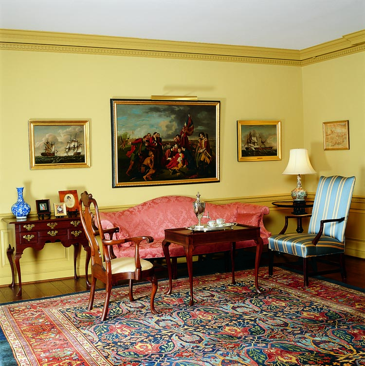 Along one end of the living room is a sitting area. The camel-back sofa is signed along the back rail by John Harris, discovered during a re-upholstery. Before purchase by its present owners the Queen Anne walnut open armchair was acquired by Wayne Pratt from the Meyer sale in 1996. The tea table exhibits the rectangular form with molded top and slipper feet recognized as among the finer from Newport, Rhode Island. The table is flanked on the other side by a Massachusetts lolling chair. The Newport dressing table along the wall, from Wayne Pratt, is one of the earliest and most dramatic examples of its type. The paintings seen here are described in the article text.